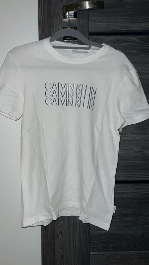 Vit Calvin Klein t-shirt med tryck - Snygg vit t-shirt från Calvin Klein med svart logotyp-tryck på bröstet. Klassisk rund hals och korta ärmar. Tillverkad i mjuk bomull som känns skön mot huden. Perfekt för dig som gillar stilrena och enkla plagg med en touch av designer.