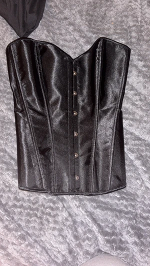 Svart korsett med snörning och hakar - Säljer en svart korsett med glansig finish och snygga metallhakar framtill. Baksidan har snörning med svarta band för att justera passformen. Korsetten har en formad siluett och ger en edgy vibe, perfekt för att lyfta din outfit.