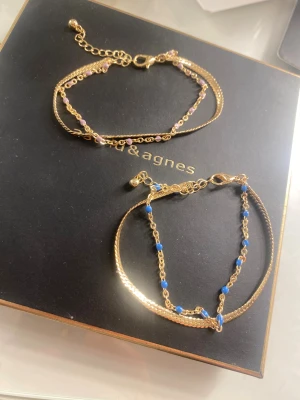 Guldarmband med färgade pärlor - Två snygga guldarmband med dubbla kedjor, ena med små blå pärlor och andra med ljusrosa pärlor. Båda har justerbar längd och smidig karbinlås. Perfekt för att ge din outfit lite extra färg och stil. Från glitter 