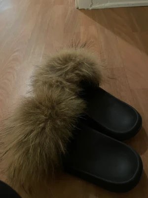 Svarta sandaler med fluffig päls - Svarta sandaler med bred rem täckt av brun fluffig päls. Snygg och chill look med platt sula och rund tå. Perfekt för dig som vill ha något extra till din outfit. Materialet är äkta päls, och färgen på pälsen är beige/brun.