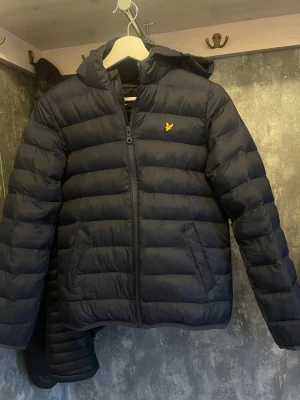 Mörkblå pufferjacka från Lyle & Scott - Snygg mörkblå pufferjacka från Lyle & Scott med huva och gul logga på bröstet. Jackan har dragkedja framtill, två sidofickor och quiltad design. Perfekt för kalla dagar och har en clean, modern look som funkar till streetwear.