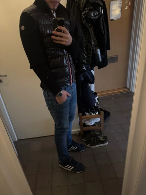 Svart stickad Moncler jacka - Snygg svart stickad jacka från Moncler med glansiga puffer-detaljer på ärmarna och klassisk logga på ena ärmen. Jackan har dragkedja och ribbad struktur, perfekt för dig som vill ha en stilren och sportig look.