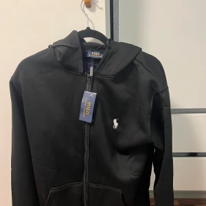 Svart hoodie från Polo Ralph Lauren - Svart hoodie från Polo Ralph Lauren med klassisk vit broderad logga på bröstet. Tröjan har huva, dragkedja framtill och två fickor. Materialet är mjuk bomull och passformen är normal. Perfekt för chill dagar och enkel att matcha med jeans eller joggers.