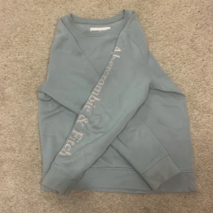 Blå sweatshirt från Abercrombie & Fitch - Snygg ljusblå sweatshirt från Abercrombie & Fitch med tryckt logga på bröstet och längs ena ärmen. Tröjan har rund hals, ribbade muddar och är tillverkad i mjuk bomullsmix. Perfekt för chill dagar och enkel att matcha med jeans eller joggers.