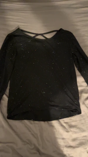 Svart glittrig topp från H&M - Svart långärmad topp från H&M med glittriga detaljer och snyggt korsade band i ryggen. Toppens material är mjukt och stretchigt, perfekt för att sticka ut. Passformen är normal och toppen har en enkel men trendig look.
