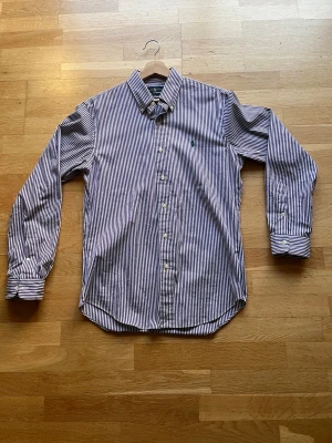 Randig skjorta från Ralph Lauren - Snygg slim fit skjorta från Ralph Lauren i vitt och mörklila med klassiskt randigt mönster. Skjortan har långa ärmar, knappar framtill och en liten broderad logga på bröstet. Perfekt för dig som gillar stilrena och tidlösa plagg.