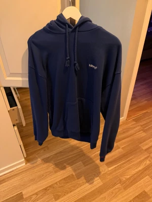 Marinblå hoodie från Levi's - Snygg marinblå hoodie från Levi's med klassisk logga på bröstet. Tröjan har huva med snörning, stor magficka och långa ärmar. Perfekt för dig som gillar en chill och avslappnad stil. Materialet är mjukt och skönt, troligtvis bomull och polyester.