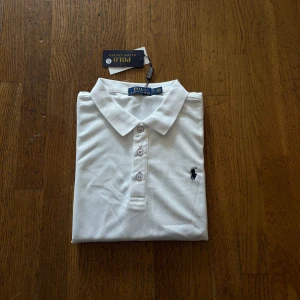 Vit Polo Ralph Lauren t-shirt - Vit t-shirt från Polo Ralph Lauren med klassisk pikékrage och tre knappar framtill. Ikoniska broderade loggan på bröstet. T-shirten är kortärmad och har en clean, stilren look som funkar till jeans eller shorts. Materialet är mjuk bomull som känns skönt mot huden.
