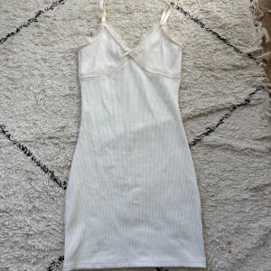 Vit ribbad kortklänning från H&M Divided - Supersöt vit ribbad klänning från H&M Divided i storlek XS. Klänningen har smala axelband, spetsdetaljer vid urringningen och en liten rosett framtill. Den är figurnära och kort, perfekt för sommarens alla tillfällen. Materialet är mjukt och stretchigt för skön passform.