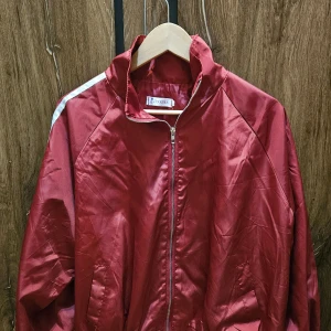 Röd vindjacka från Panpan med stripes - Röd vindjacka från Panpan med vita dubbla stripes längs ärmarna. Jackan har dragkedja framtill, hög krage och resår i midjan. Tillverkad i glansigt syntetmaterial, perfekt för en sportig och chill look.