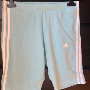 Ljusblå/turkos Adidas shorts med vita ränder - Snygga ljusblå shorts från Adidas med klassiska vita ränder längs sidorna och Adidas-logga framtill. Mjukt material och elastisk midja för skön passform. Perfekta för träning eller chill dagar. Passar dig som gillar sportig stil.