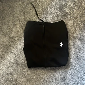 Svart hoodie från Polo Ralph Lauren - Svart hoodie från Polo Ralph Lauren med dragkedja och huva med snörning. Klassisk vit logga broderad på bröstet och två fickor framtill. Mjuk och skön, perfekt för chill dagar. Långärmad och i bomullsmix.
