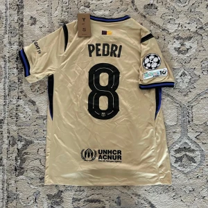 FC Barcelona Pedri #8 Champions League tröja - Champagnefärgad FC Barcelona fotbollströja från Nike med Pedri och nummer 8 på ryggen. Champions League-märke på ärmen, svarta detaljer och blå inslag vid krage och ärmslut. Materialet är lätt och ventilerande polyester, perfekt för match eller träning.