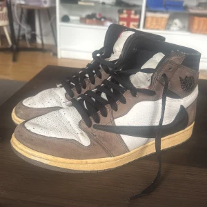 Nike Air Jordan 1 Travis Scott High - Nike Air Jordan 1 Travis Scott High med bruna mockadetaljer, vit läderbas och svarta snören. Swoosh-loggan är svart och vänd åt fel håll på utsidan. Gul vintage-inspirerad sula och diskret Cactus Jack-logga på hälen. Perfekta för dig som vill sticka ut. Tecken på användning därav priset. Låda och allt OG följer med, från Monicasneaker