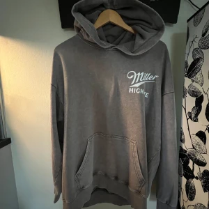 Grå hoodie Miller High Life L - Snygg grå hoodie med stor huva och tryck både fram och bak från Miller High Life. Oversized passform, känguruficka och coolt retroinspirerat tryck med texten 'The Champagne of Beers'. Perfekt för chill dagar och streetstyle.
