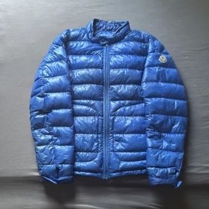 Blå Moncler dunjacka med dragkedja - Säljer en blå dunjacka från Moncler med glansig finish och klassisk logga på ärmen. Jackan har hög krage, dragkedja framtill och quiltad design. Perfekt för kalla dagar när du vill ha både stil och värme.