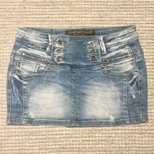 Blå jeanskjol från Dromedar - Snygg blå jeanskjol från Dromedar med coola detaljer som knappar och dragkedjor framtill. Kjolen har en kort längd och två bakfickor. Perfekt för en avslappnad stil. Står att den e i storlek 29 men skulle verkligen säga mer en xs-s! Detta märks även på måtten som finns på bild 4 och 5 (midjemått 37 cm tvärsöver o längd framtill 27 cm) 🩷