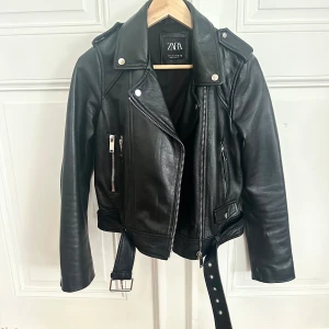 Skinnjacka från Zara - Snygg äkta svart skinnjacka från Zara med klassisk bikerstil, silverfärgade dragkedjor och bälte i midjan. Jackan har långa ärmar, bred krage och coola detaljer som axelklaffar och flera fickor. Perfekt för dig som vill ha en edgy look. Väldigt lite tecken på användning då jag använt den ett fåtal ggr.
