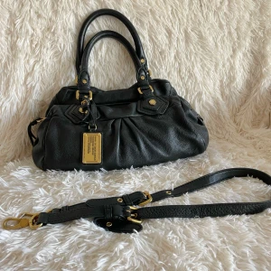 Svart handväska Marc by Marc Jacobs - Snygg svart handväska i skinn från Marc by Marc Jacobs med guldfärgade metalldetaljer och avtagbar axelrem. Väskan har dubbla handtag, dekorativa sömmar och en stor metallbricka framtill. Invändigt foder med logotypmönster och dragkedja upptill. Storlek: 30cm x 20cm x 10cm