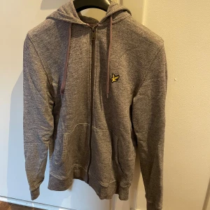 Grå hoodie med dragkedja från Lyle & Scott - Gråmelerad hoodie från Lyle & Scott med gul logga på bröstet. Tröjan har huva med snörning, hel dragkedja och två fickor framtill. Perfekt för chill dagar och enkel att matcha med jeans eller joggers.