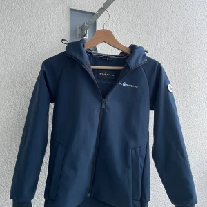 Marinblå vindjacka från Sail Racing - Snygg marinblå vindjacka från Sail Racing med huva och dragkedja. Jackan har en stilren design med logga på bröstet och är fodrad med mjuk fleece på insidan för extra komfort. Perfekt för blåsiga dagar och enkel att matcha med det mesta.