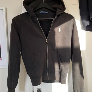 Svart hoodie från Polo Ralph Lauren - Svart hoodie från Polo Ralph Lauren med vit broderad logga på bröstet. Tröjan har dragkedja framtill, huva och två fickor. Tillverkad i mjuk bomull och har långa ärmar samt ribbade muddar för en skön passform