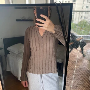 Brun ribbad långärmad topp - Säljer en brun långärmad topp med ribbat, vertikalt mönster och v-ringning med knappar framtill. Tunt och stretchigt material som sitter snyggt på kroppen. Perfekt att styla med jeans eller kjol för en chill look.