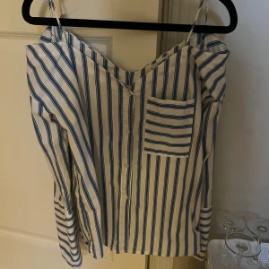 Randig blus med smala axelband - Snygg blus med blå och vita ränder, smala axelband och v-ringning. Kan andvändas med de små banden eller som offshoulder. Passar både m och s 