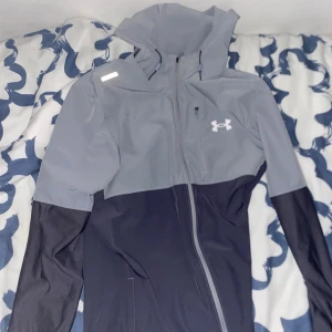 Grå och svart vindjacka Under Armour (äkta) - Snygg vindjacka från Under Armour i grått och svart med huva och hel dragkedja. Jackan har en sportig look, logga på bröstet och är tillverkad i lätt polyester. Perfekt för dig som gillar att vara aktiv och vill ha en stilren jacka med bra passform.