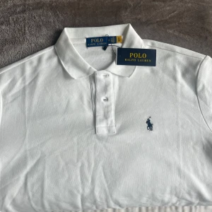 Vit Polo Ralph Lauren piké M - Vit klassisk t-shirt från Polo Ralph Lauren med korta ärmar och krage. Ikonisk broderad logga i mörkblått på bröstet. Tillverkad i mjuk bomull som känns skön mot huden. Perfekt för dig som gillar stilrena och tidlösa plagg.