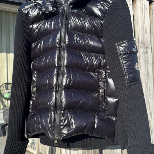 Svart Moncler cardigan  - Snygg svart cardigan från Moncler med glansig front och luva, samt stickade ärmar och rygg. Jackan har dragkedja framtill, fickor med tryckknappar och en cool ärmficka med Moncler-logga. Perfekt för dig som vill ha en stilren och exklusiv look. Hör av dig vid frågor!