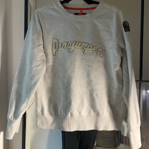 Ljusgrå fleece-tröja från Parajumpers - Jag säljer en ljusgrå fleeceskjorta från Parajumpers med en broderad logotyp på bröstet och ett märke på ena ärmen. Skjortan har rund halsringning, långa ärmar och ribbade muddar. Perfekt för kalla dagar och riktigt bekväm att bära. Passar storlekar S-L.