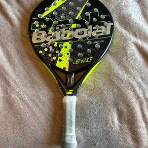 Defiance - Babolat Defiance är ett padelracket med modern design och coola färger. Perfekt för dig som vill ta ditt spel till nästa nivå, oavsett om du är nybörjare eller mer erfaren. Racketen väger 350g och har en balans på 265mm, vilket ger bra kontroll och känsla på banan.