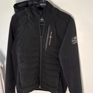 Svart pufferjacka från Sail Racing - Svart pufferjacka från Sail Racing med huva och dragkedja framtill. Jackan har quiltad framsida, diskret logga på bröstet och ärm, samt en snygg, sportig look. Perfekt för dig som gillar stilren och funktionell design.