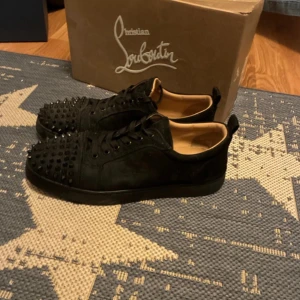 Christian Louboutin svarta sneakers med nitar - Svarta sneakers från Christian Louboutin i mocka med coola svarta nitar på tån och klassisk röd sula. Skorna har snörning och en rund tå, vilket ger en edgy och lyxig vibe. Perfekta för dig som vill sticka ut med exklusiva detaljer.