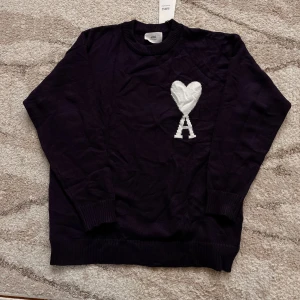Mörkblå stickad tröja från Ami - Säljer en mörkblå stickad tröja från Ami med stor vit hjärtlogga och bokstaven A på bröstet. Tröjan har rund hals, ribbade muddar och lång ärm. Perfekt för dig som gillar stilrena plagg med en twist.