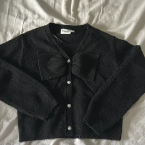 Svart kofta med rosett från Kappahl - Supersöt svart kofta från Kappahl i stickat material. Koftan har långa ärmar, pärlknappar framtill och en stor rosett vid bröstet som ger en extra touch. Perfekt för dig som gillar stilrena men ändå unika plagg. Aldrig använt!