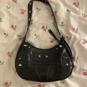 Svart croco axelväska med nitar - Svart axelväska i croco-mönstrat syntetmaterial med silvriga nitar och detaljer. Väskan har justerbart axelband med spänne och dragkedja upptill. Insidan har flera fack för att hålla ordning på dina grejer. 