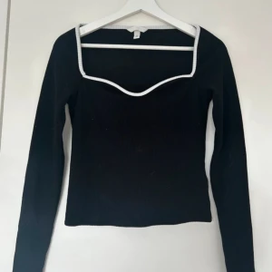 Svart ribbad topp med vit kant - Snygg svart ribbad topp med lång ärm och vit kant runt den fyrkantiga urringningen. Toppen har en figurnära passform och är perfekt för att lyfta en enkel outfit. Materialet är stretchigt och bekvämt.