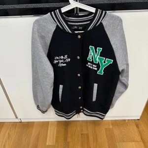 Svart och grå bomberjacka NY - Snygg svart och grå bomberjacka från LGV med broderad NY-logga i grönt och vitt på bröstet. Jackan har grå ärmar, svarta muddar med vita ränder och knappar framtill.