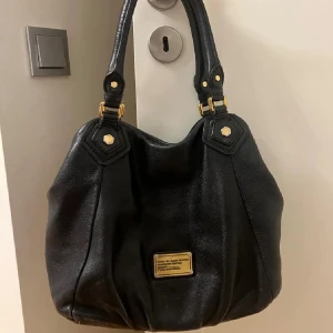 Marc Jacobs väska - Säljer en svart axelväska i skinn från Marc by Marc Jacobs med guldiga metalldetaljer och en snygg logoplatta framtill. Väskan har dubbla handtag och ett rymligt innerfack med coolt mönstrat foder med märkets logga. Perfekt för dig som vill ha en stilren och praktisk väska.