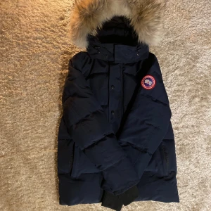 Canada Goose Wyndham Parka  - Säljer en mörkblå Canada Goose dunjacka med stor fluffig pälskantad huva och klassisk logga på ärmen. Jackan har dragkedja och knappar framtill, två stora fickor och ribbade muddar vid ärmsluten. Perfekt för kalla vinterdagar. Väldigt varm och vattentät.
