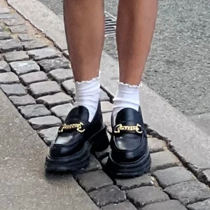 Chunky svarta loafers med guldkedja - Stilrena svarta loafers i skinn med chunky platt sula och dekorativ guldfärgad kedjedetalj framtill. Perfekta för att lyfta din outfit med en trendig touch och funkar lika bra till jeans som kjol.