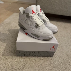 Nike Air Jordan 4 vit/grå sneakers - Nike Air Jordan 4 sneakers i vitt och grått med röda detaljer. Skorna har klassisk snörning, meshpaneler och en platt sula med synlig Air-enhet. Ikonisk Jumpman-logga på plös och sula. Perfekt för dig som gillar streetstyle och vill sticka ut med en clean look.