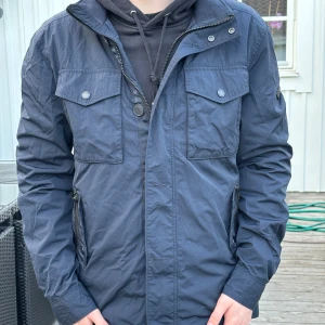 Blå vindjacka från J.Lindeberg - Snygg mörkblå vindjacka från J.Lindeberg med dragkedja och knappar framtill. Perfekt nu till hösten och kallare dagar. Den är i väldigt bra skick och har mycket mer. Jackan är Storlek S, men stor i storleken.