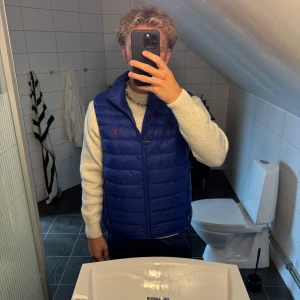 Blå dunväst från Polo Ralph Lauren - Snygg blå dunväst från Polo Ralph Lauren med klassisk röd logga på bröstet. Västen har quiltad design, dragkedja framtill och två fickor med dragkedja. Perfekt att slänga över en hoodie eller tröja för en sportig och stilren look.