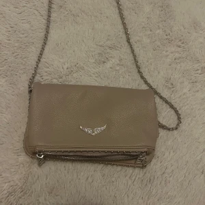 Beige Zadig& Voltaire axelväska - Snygg beige Zadig & Voltaire axelväska i skinnimitation med silverfärgad kedjerem och dekorativt emblem. Bilderna gör inte färgen på väskan faire utan den är med rosa beige i verkligheten. Köpt 2 år sedan 