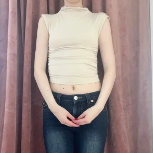 Beige croppad ärmlös topp - Säljer en beige croppad topp med hög hals och ärmlös design. Toppen har en tight passform och snygga rynkade detaljer i sidorna 