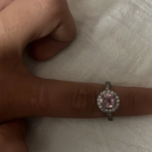 Silverring med rosa sten och glittriga detaljer - Snygg silverfärgad ring med en rund rosa sten i mitten, omgiven av små glittriga stenar som ger extra bling. Ringen har en klassisk form och passar perfekt om du vill sticka ut med något unikt och elegant. Perfekt för dig som gillar färg och glitter! Nästan helt oanvänd 