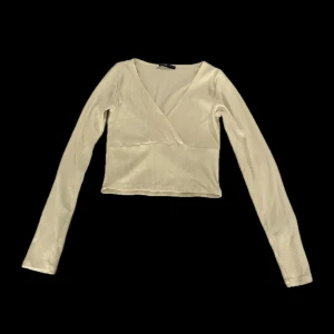 Crèmefärgad ribbad omlottopp med v-ringning - Snygg Crèmefärgad långärmad topp med ribbad struktur och omlott-design framtill. Toppen har en djup v-ringning och croppad passform som ger en trendig look. Perfekt att ha ett linne under. Köpt på newyorker.